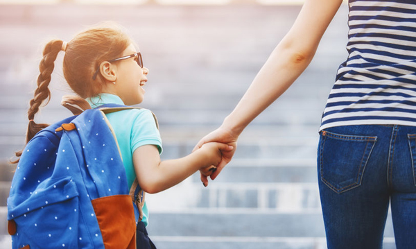 A young girl holds her mother's hand as they walk together. She carries a backpack on her shoulder.