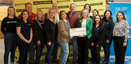 Fourteen people stand together, including members of the team from Reading is Fundamental and Dollar General. Two people hold an oversized check in the center of the group.