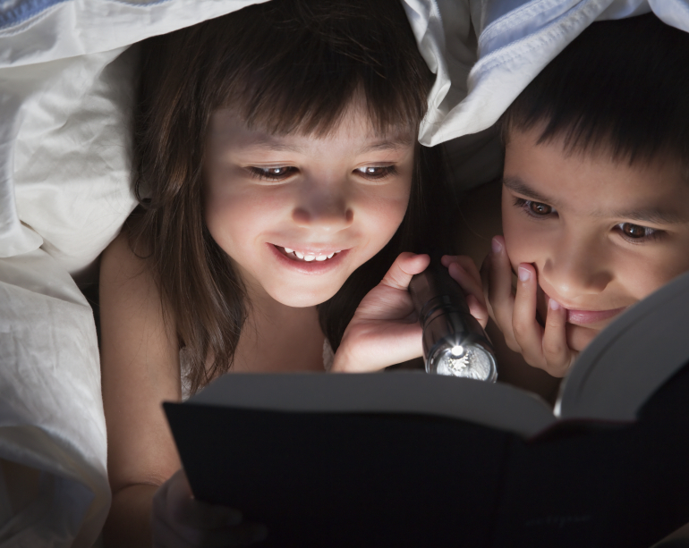 Kids smiling happy reading a book under a blanket with flashlight