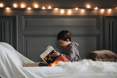 A boy in a gray jacket reads a book on his bed. Shining string lights are hung from the wall above him.