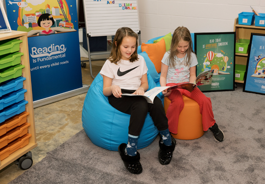 Two girls read in the new library created with the help of RIF and Dollar General. They are seated in bean bag chairs beside each other with books open. 
