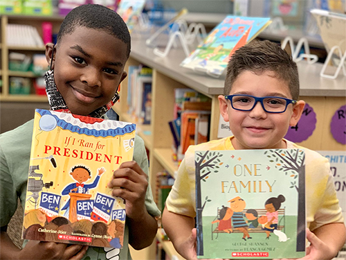 Two boys stand together, each holding a book from a RIF event. The boy on the left holds "If I Ran for President" and the boy on the right holds "One Family".