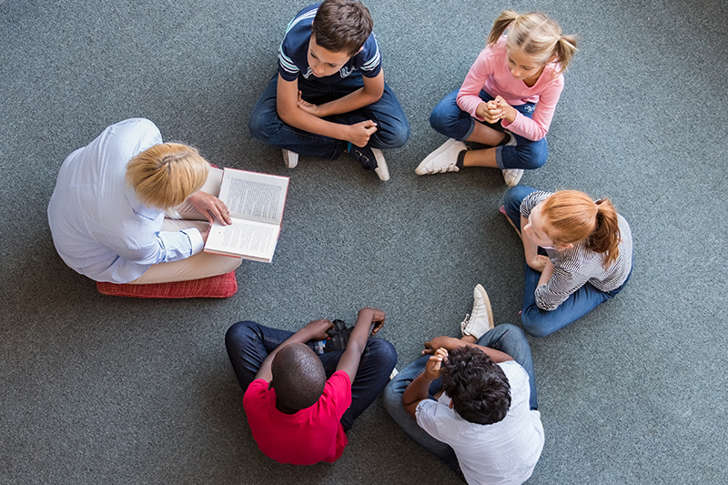 A group of children are seated in a circle while they read a book together