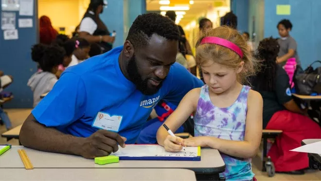 An image of an Amazon employee wearing a blue shirt with the company logo helping a student write something down on a white pad. The student is wearing a tie-dye shirt and a pink headband.  